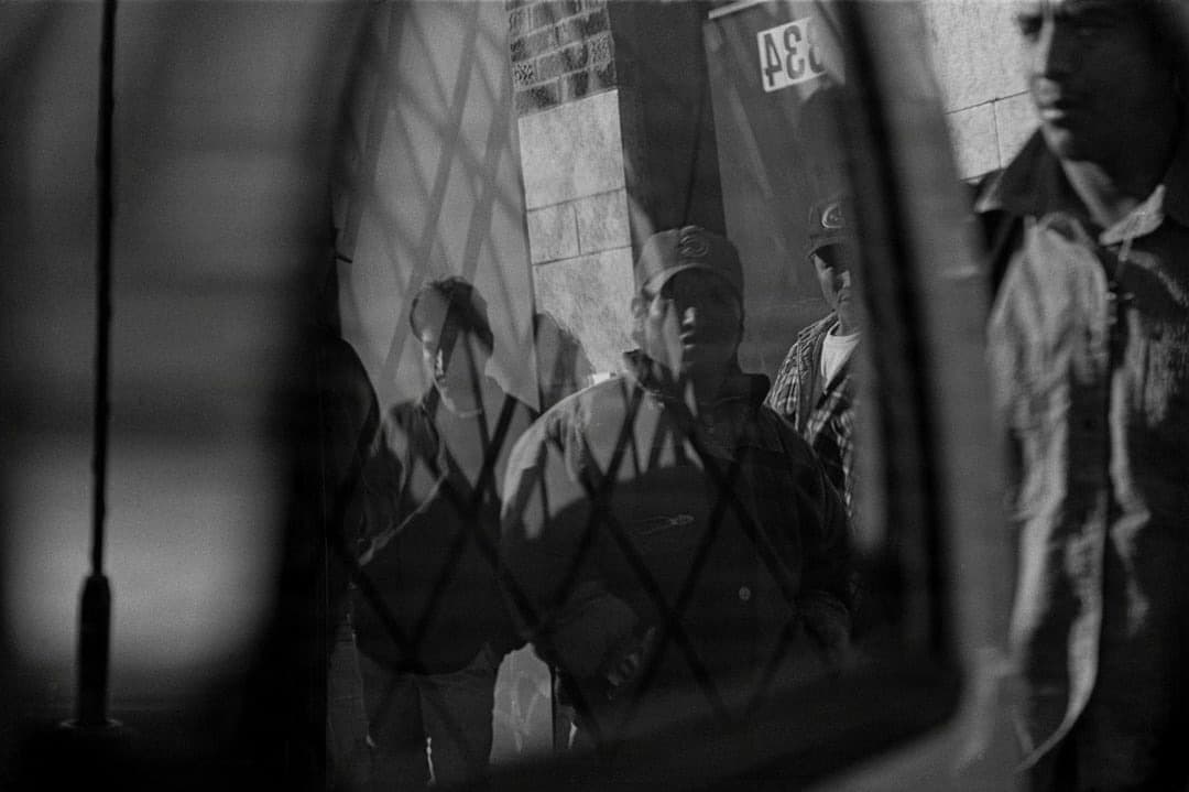 Urban figures observed through a patterned window, their faces partially obscured.
