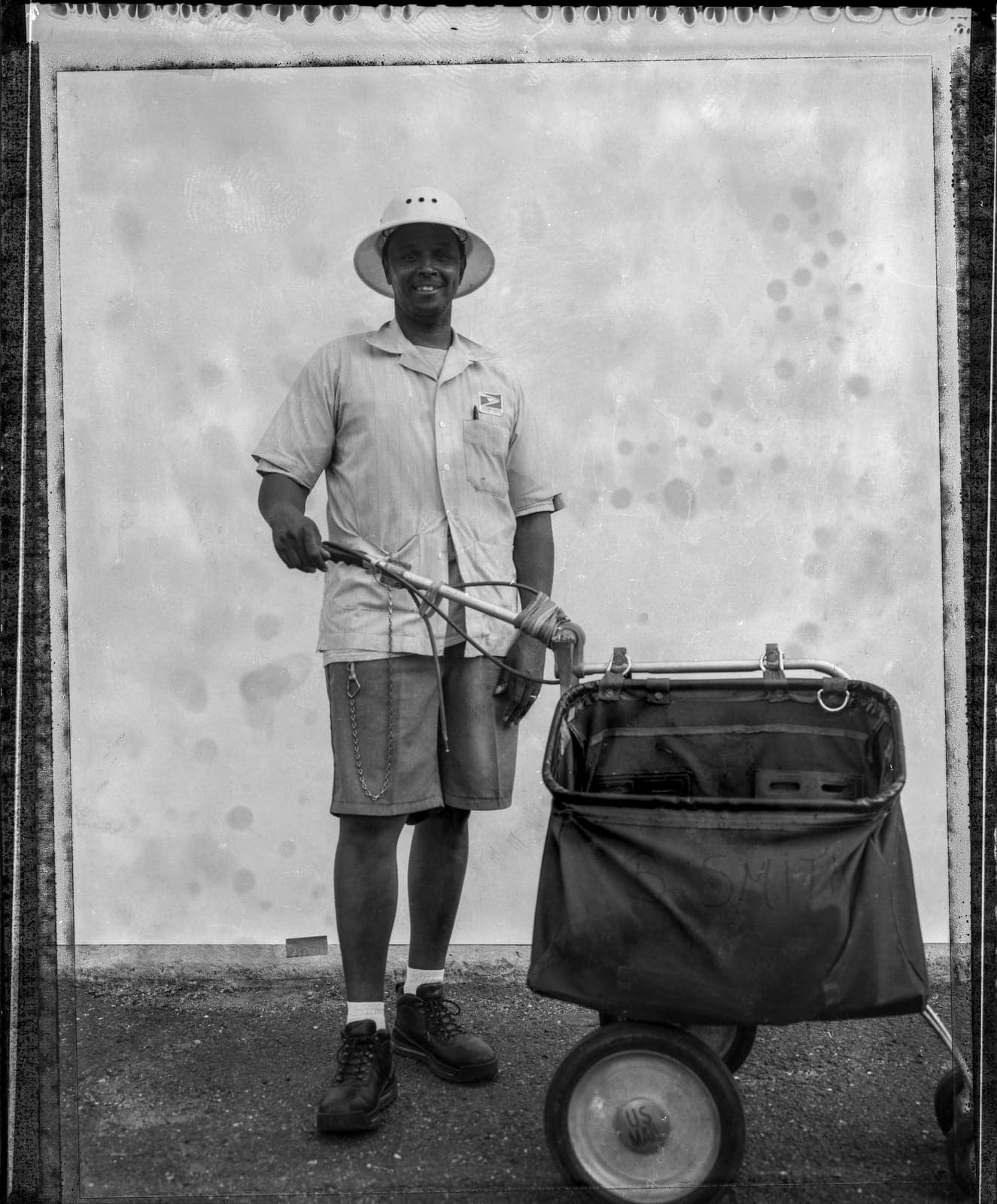 Mail carrier in uniform with pith helmet stands beside pushcart.